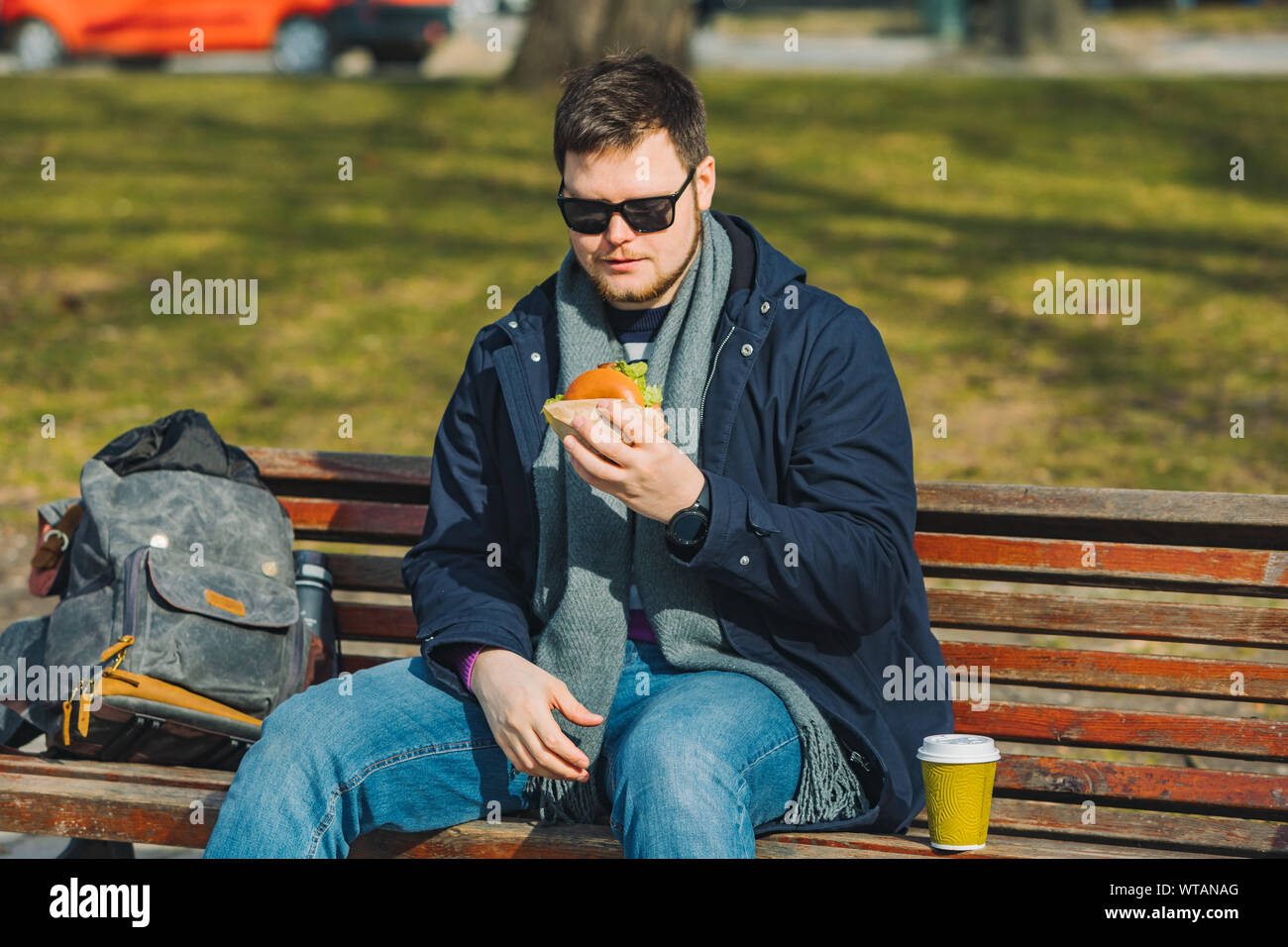 young smiling man eating burger and drink coffee sitting on city bench Stock Photo