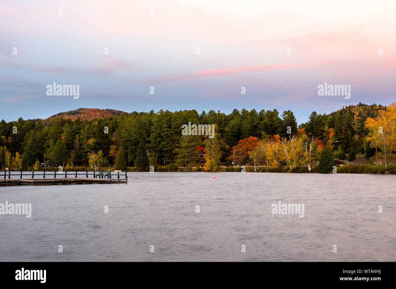 Lake surrounded by colourful autumn trees at twilight. A deserted wooden jetty is visible in foreground. Stock Photo