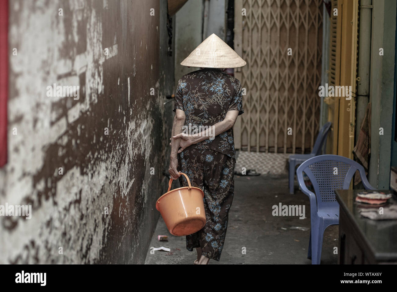 Old woman wears Nao La hat carrying a bucket in Saigon alleys Stock Photo