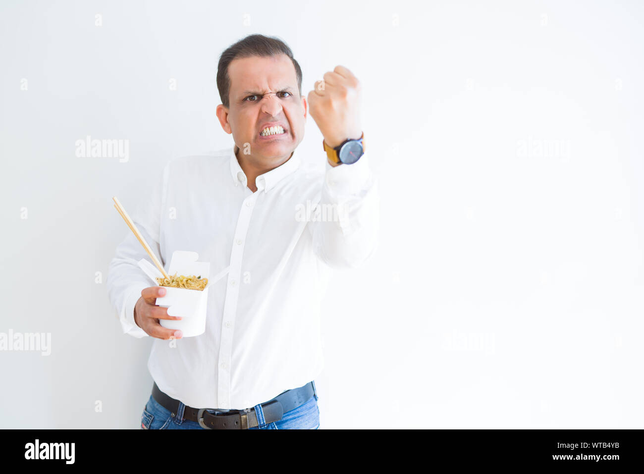 Middle age man eating asian noodles from take away carton annoyed and frustrated shouting with anger, crazy and yelling with raised hand, anger concep Stock Photo