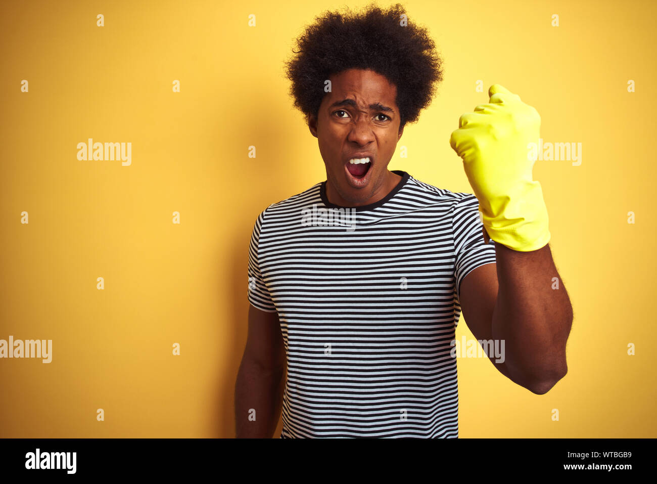 Young african american man cleaning using gloves standing over isolated yellow background annoyed and frustrated shouting with anger, crazy and yellin Stock Photo