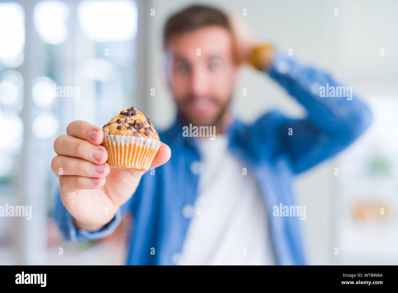 Handsome man eating chocolate chips muffin stressed with hand on head