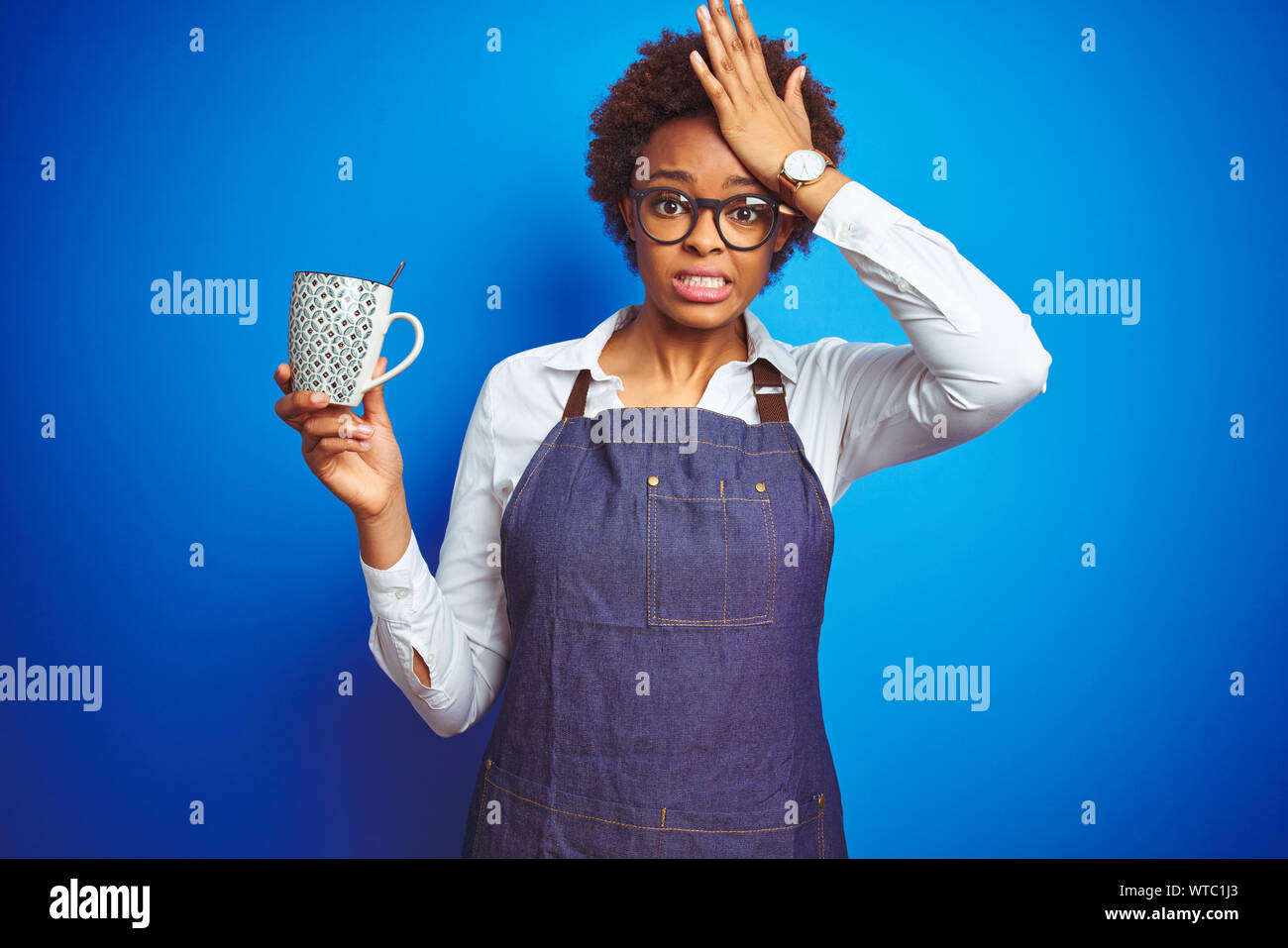 African american barista woman wearing bartender uniform holding cup over blue background stressed with hand on head, shocked with shame and surprise Stock Photo