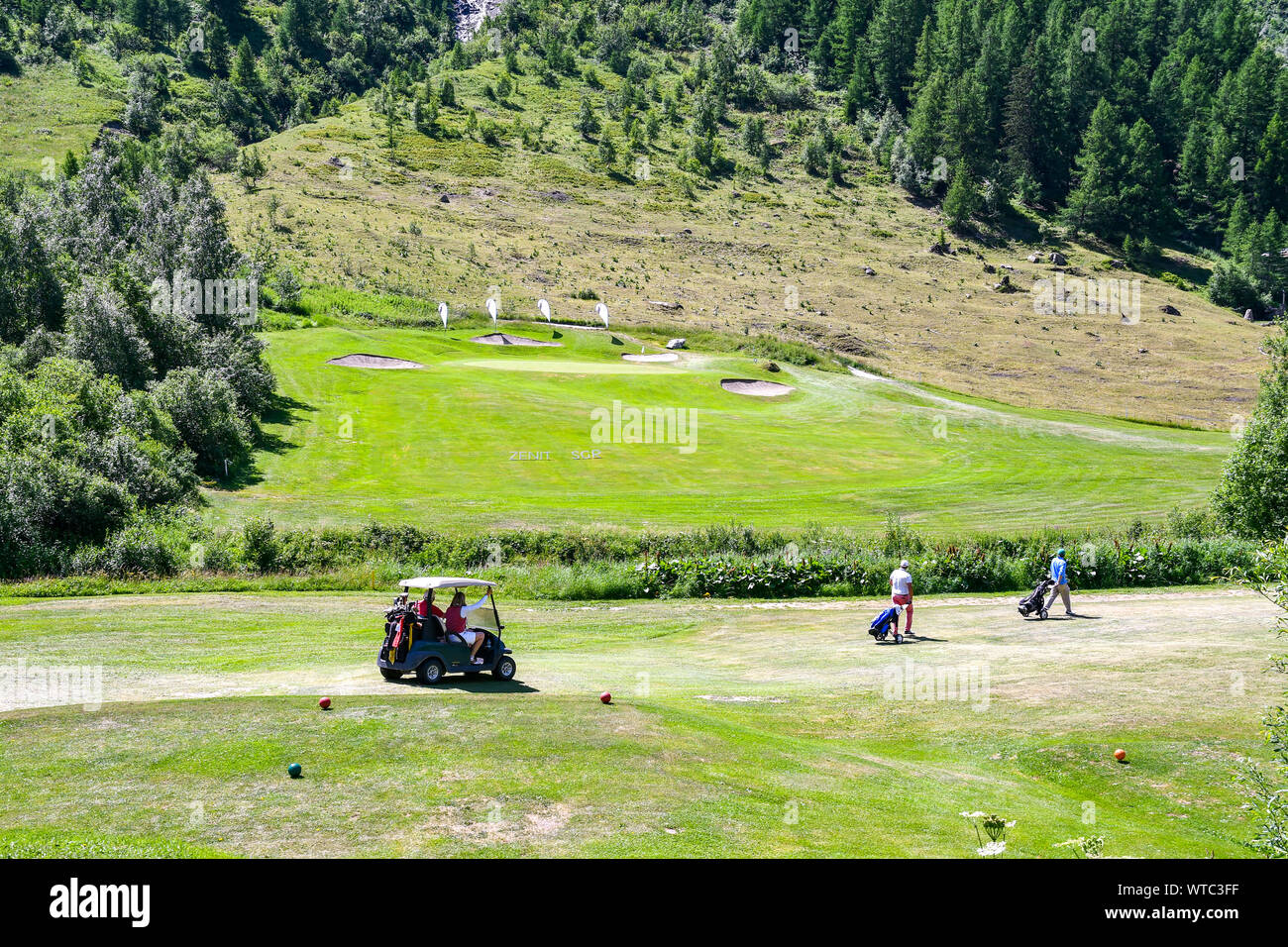 Scenic view of a mountain golf course with middle-aged people on a golf cart and carrying golf bags, Val Ferret, Courmayeur, Aosta Valley, Alps, Italy Stock Photo