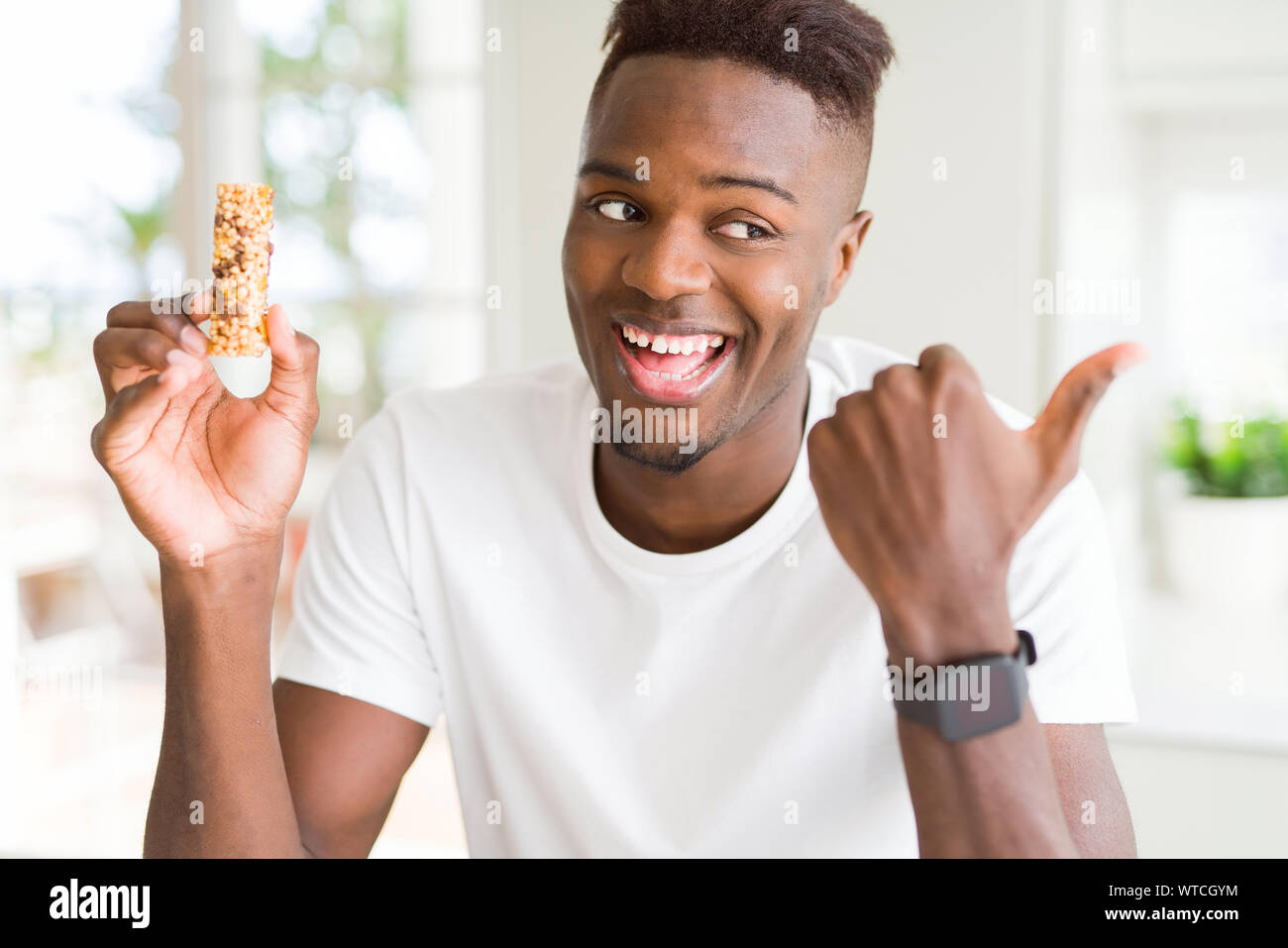 African american man eating energetic cereals bar pointing and showing with  thumb up to the side with happy face smiling Stock Photo - Alamy, image size:1300x957