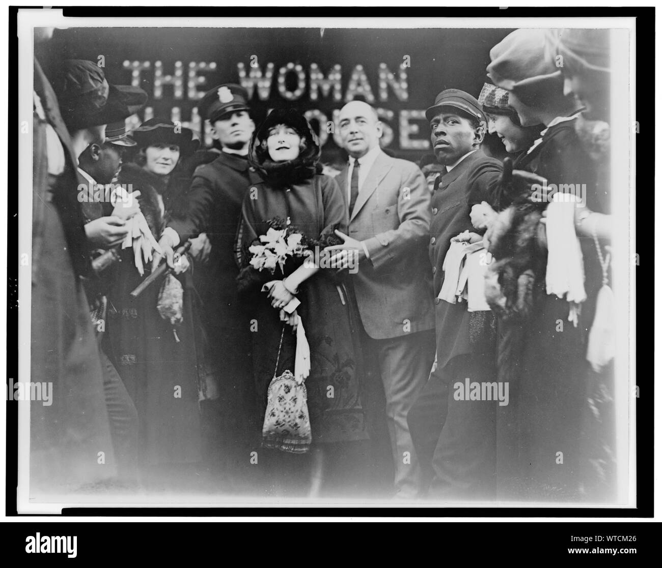 Mildred Harris Chaplin, full-length portrait, standing in crowd, facing front, guarded by a policeman, at a theater showing her film The Woman in His House (1920). Stock Photo