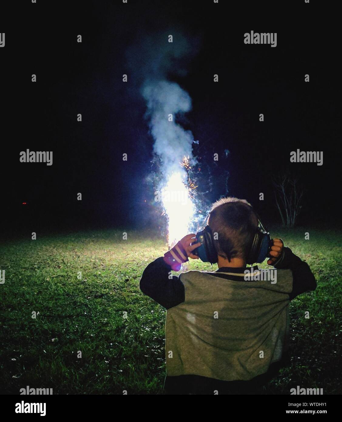 Rear View Of Boy Wearing Ear Protectors Standing On Field With Firework At Night Stock Photo