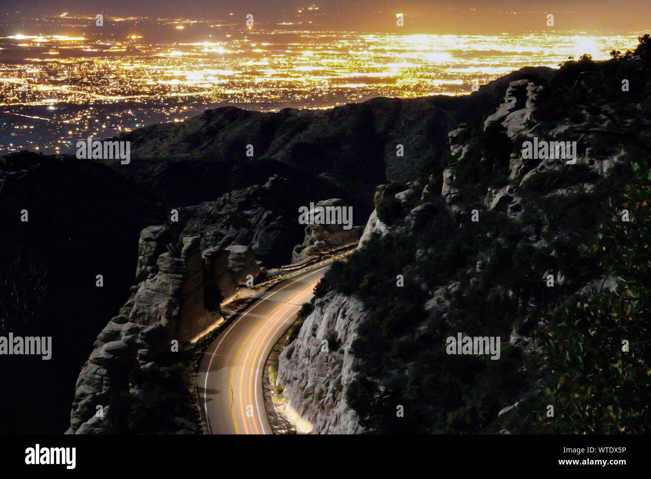Light Trail On Mountain Road Against Illuminated City Stock Photo