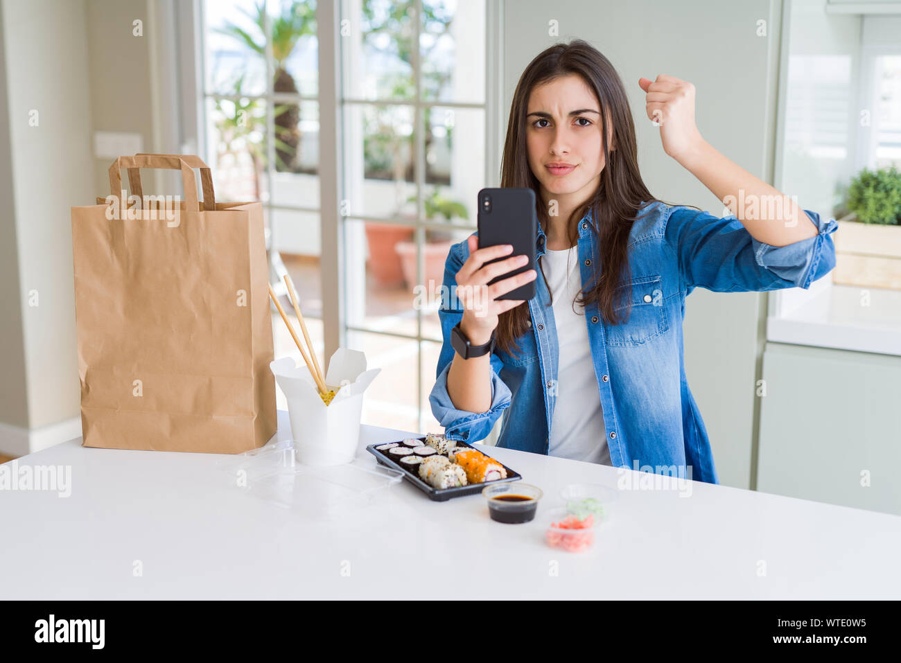 Beautiful young woman ordering food delivery from app using smartphone annoyed and frustrated shouting with anger, crazy and yelling with raised hand, Stock Photo