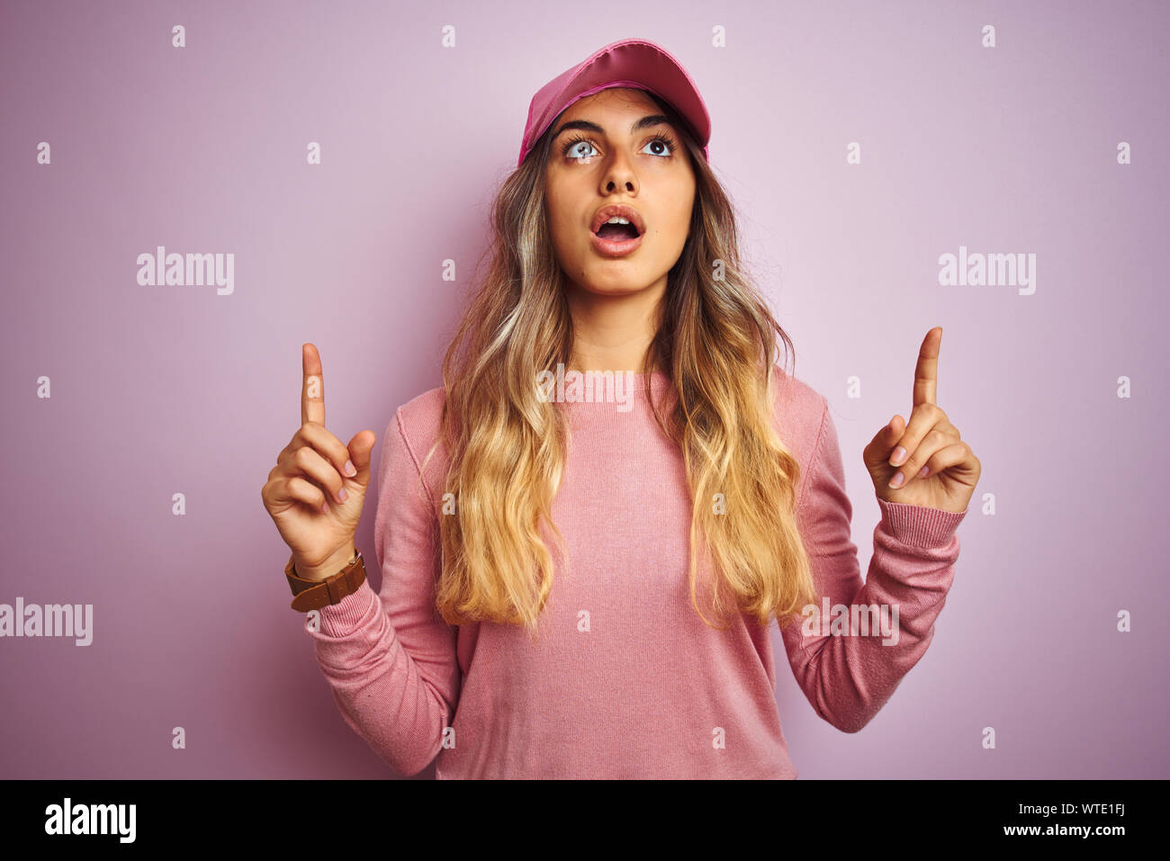 Young beautiful woman wearing cap over pink isolated background amazed and surprised looking up and pointing with fingers and raised arms. Stock Photo