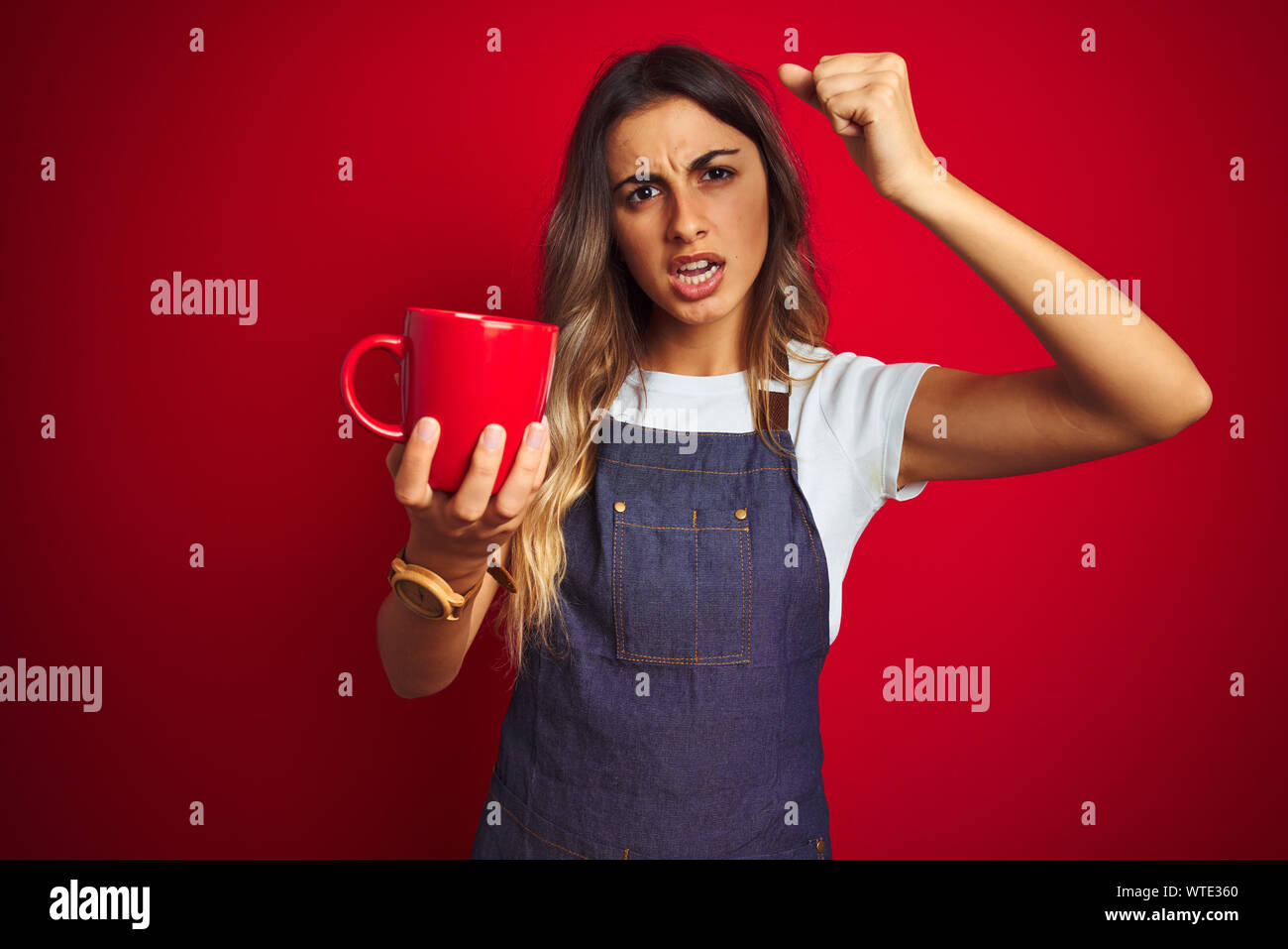 Young beautiful barista woman wearing apron over red isolated background annoyed and frustrated shouting with anger, crazy and yelling with raised han Stock Photo