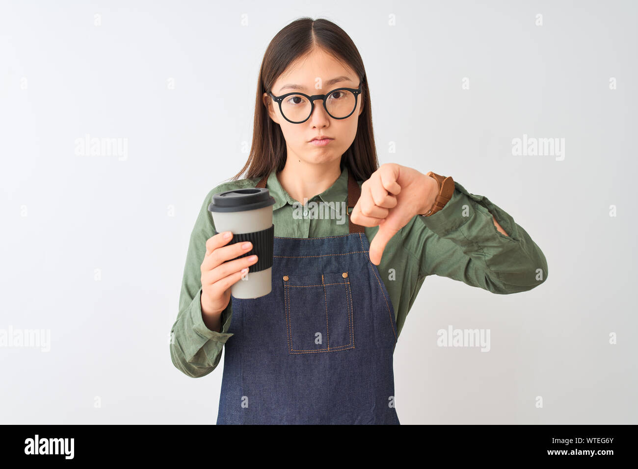 Chinese barista woman wearing apron glasses drinking coffee over isolated white background with angry face, negative sign showing dislike with thumbs Stock Photo