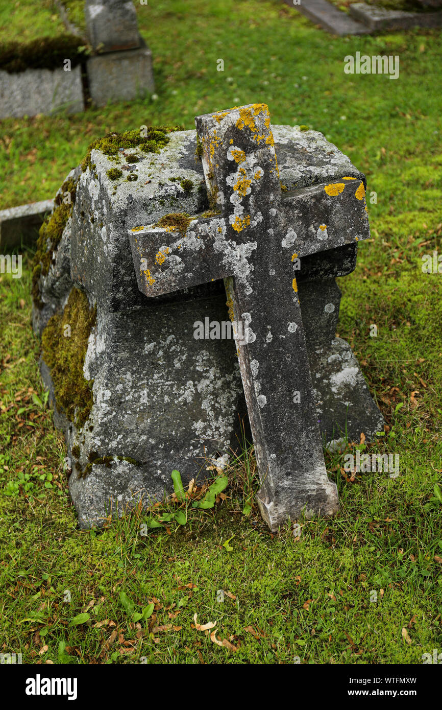 Fallen cross, covered with lichen and moss, leaning against headstone Stock Photo