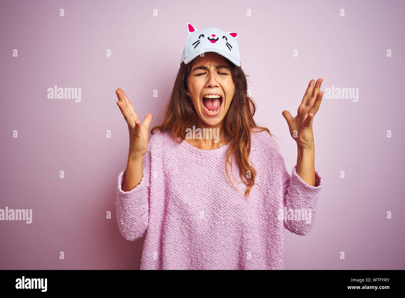 Young beautiful woman wearing funny cat cap standing over pink isolated background celebrating mad and crazy for success with arms raised and closed e Stock Photo