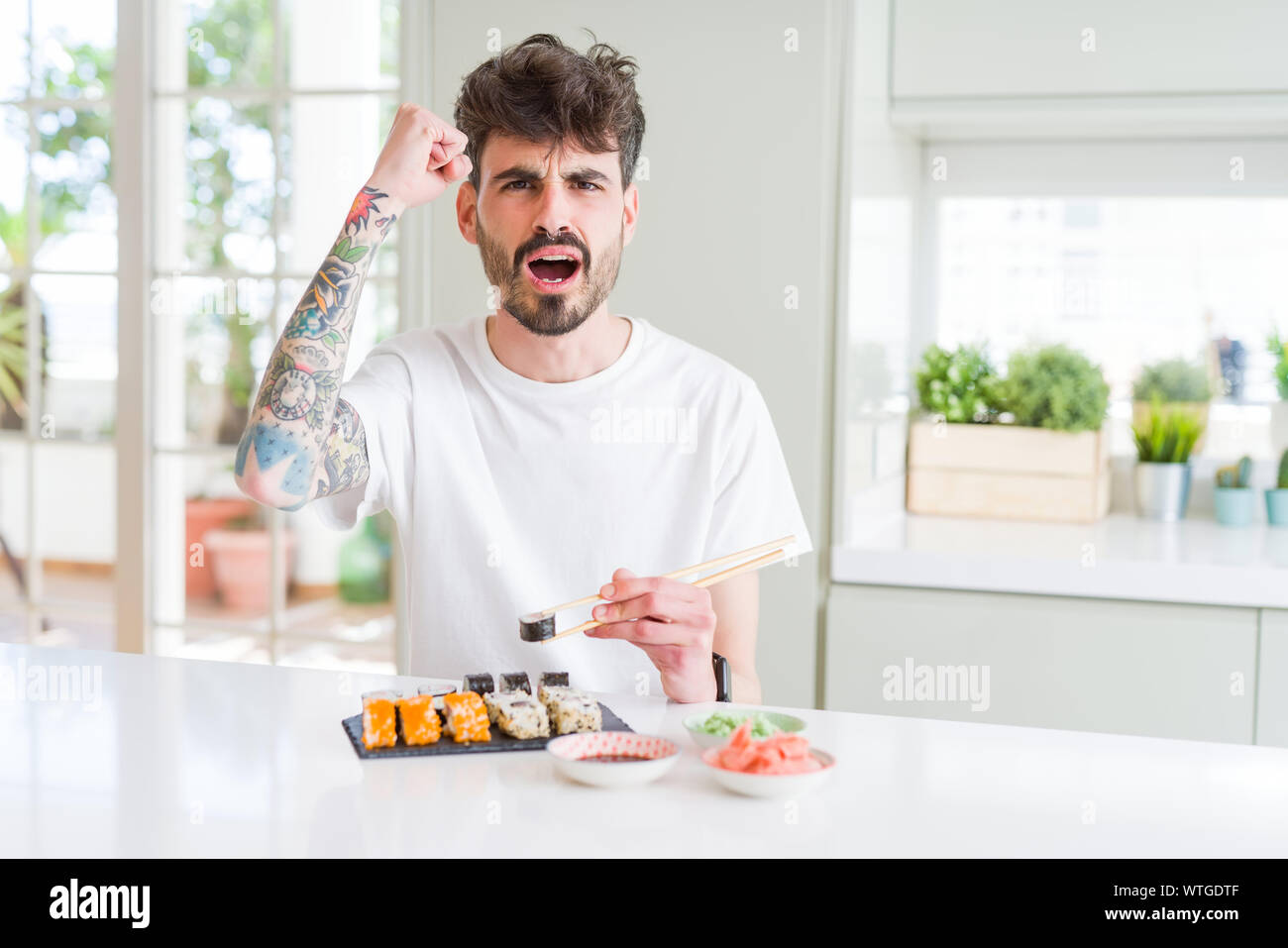 Young man eating asian sushi from home delivery annoyed and frustrated shouting with anger, crazy and yelling with raised hand, anger concept Stock Photo