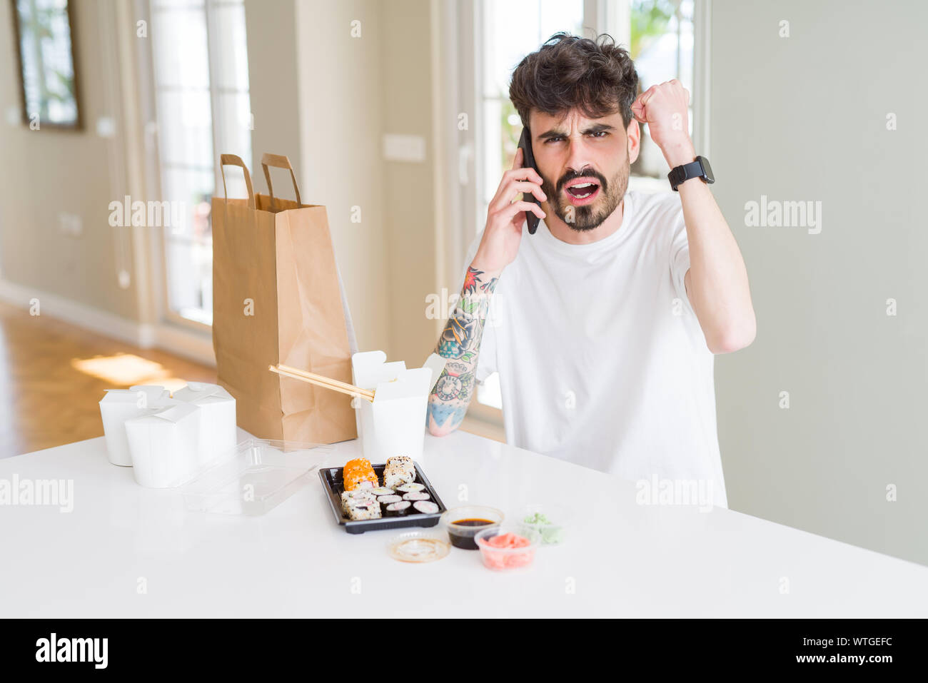 Young man eating asian sushi from home delivery and ordering food talking on smartphone annoyed and frustrated shouting with anger, crazy and yelling Stock Photo
