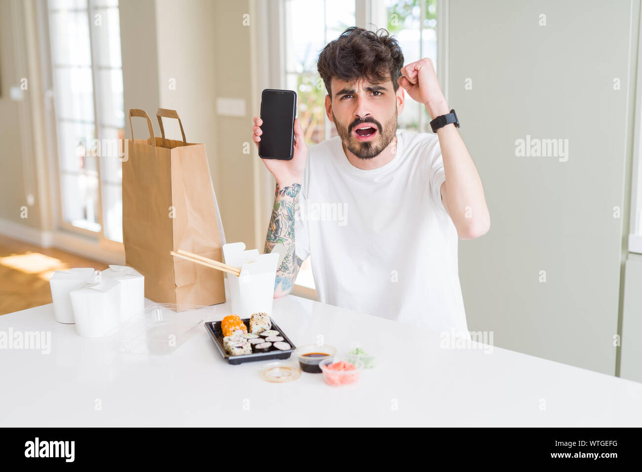 Young man eating asian sushi from home delivery and showing smartphone screen annoyed and frustrated shouting with anger, crazy and yelling with raise Stock Photo