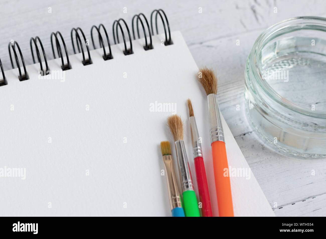 Art pad, paint brushes and glass of water on a wooden surface. Artist desk with a very shallow depth of field Stock Photo