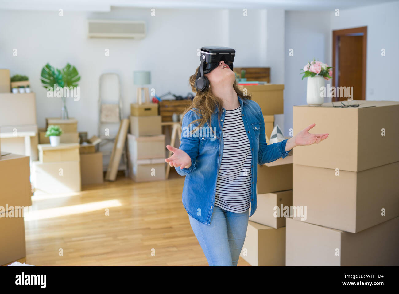 Young blonde woman wearing virtual reality glasses playing a simulation game around cardboard boxes moving to a new house Stock Photo