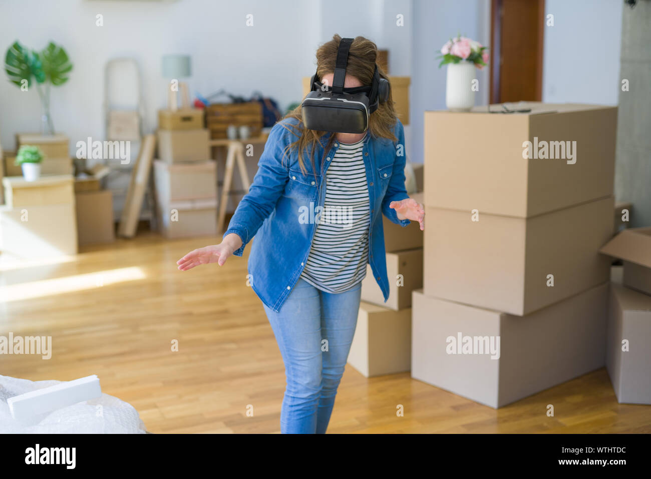 Young blonde woman wearing virtual reality glasses playing a simulation game around cardboard boxes moving to a new house Stock Photo