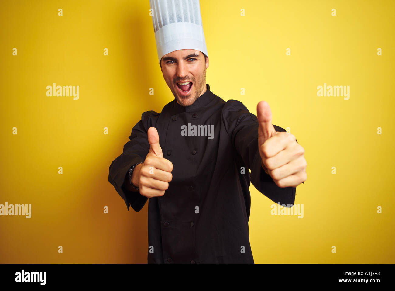 Young chef man wearing uniform and hat standing over isolated yellow background approving doing positive gesture with hand, thumbs up smiling and happ Stock Photo