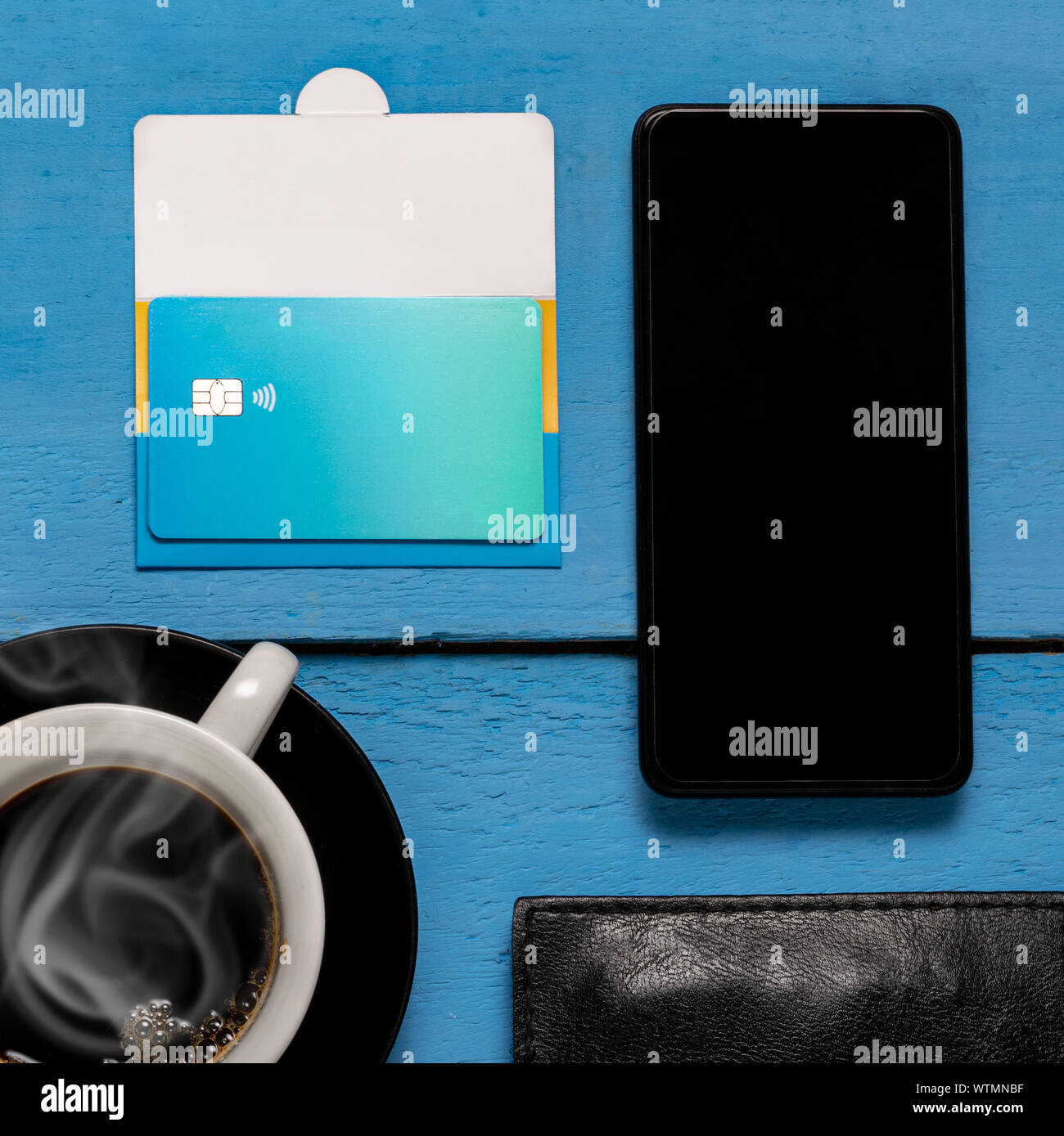 A blue contactless credit card showing chip, with a smartphone, coffee cup and leather wallet on top of a blue wooden background Stock Photo