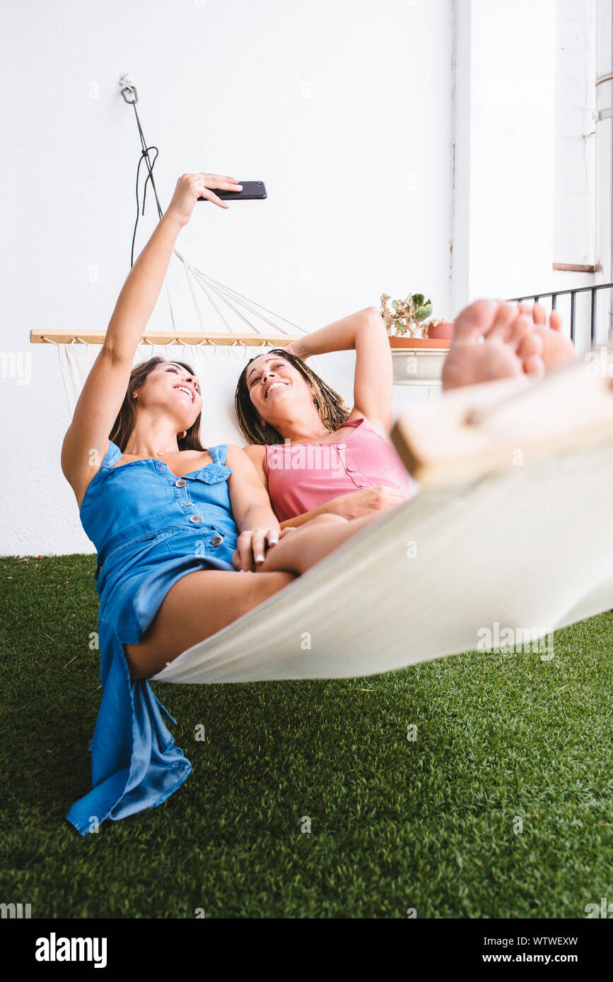 Two barefoot women lying on hammock taking a selfie. Stock Photo