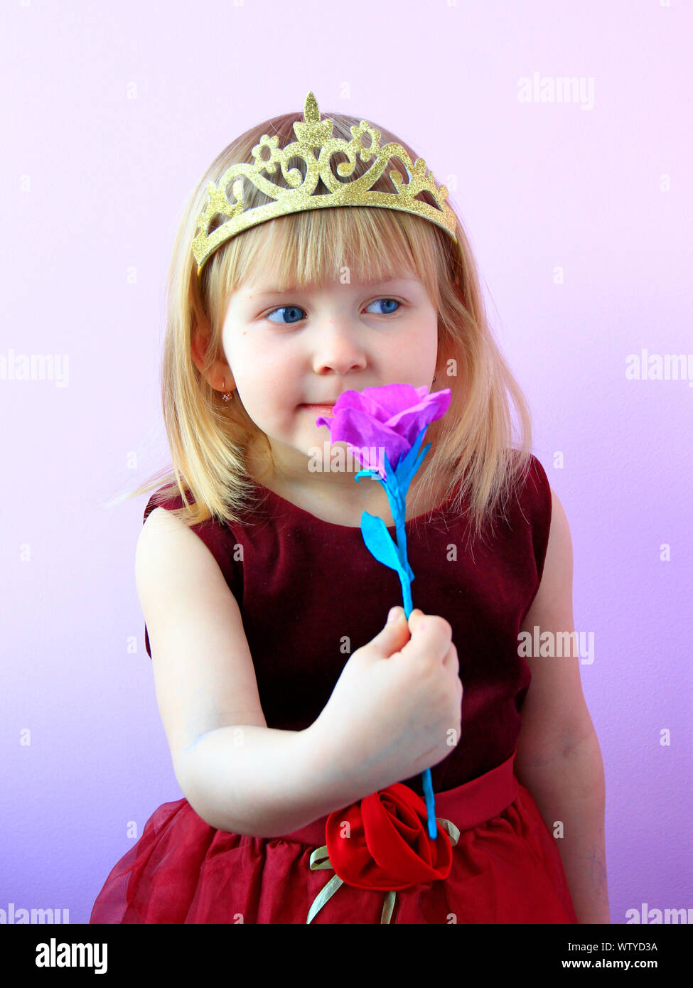 Little girl in crown smelling flower. Little princess with pink flower. Cute girl with crown on head offering pink rose. Child in beautiful dress smil Stock Photo