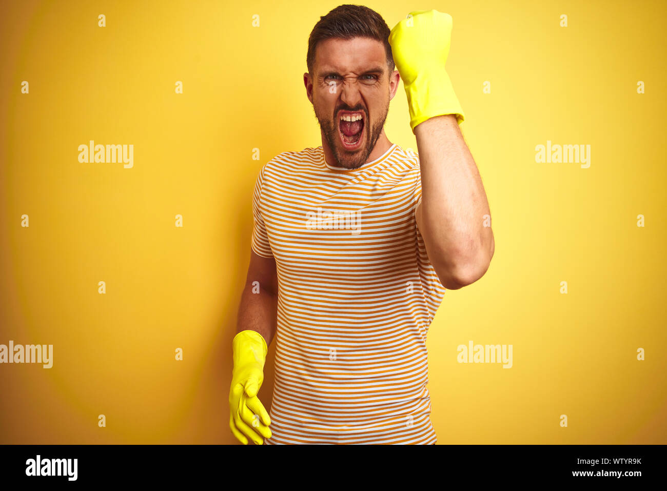 Young handsome man wearing cleaning gloves for housework over isolated yellow background annoyed and frustrated shouting with anger, crazy and yelling Stock Photo