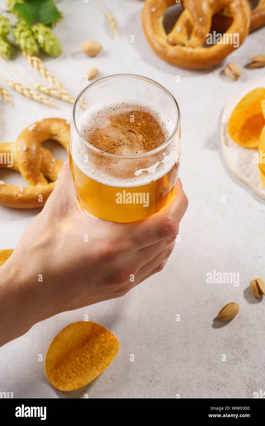 Man hand holding a glass of light beer. Various snacks in the background - pretzels, chips and nuts. Stock Photo