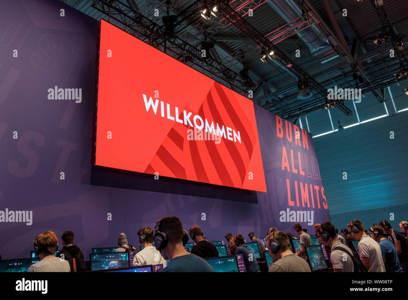 visitors play car racing game  below large screen displaying the word „Willkommen“ (German for „welcome“) at gamescom , world’s largest trade fair for computer and video games in Cologne, Germany on  August 21, 2019 Stock Photo