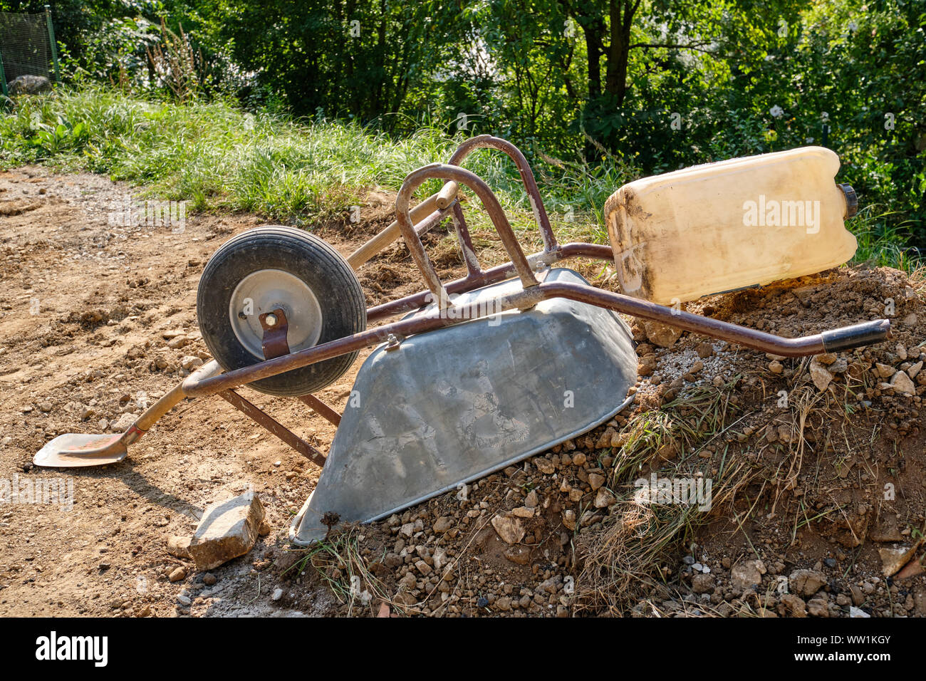 A wheelbarrow is lying upside down on a heap of soil and stones together with a shovel and a canister on a small construction site in Germany Stock Photo