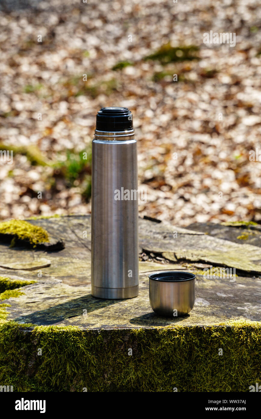 Thermos bottle and a cup of coffee or tea on a moss covered tree stump in sunlight in the woods (eye-level view, vertical format) Stock Photo