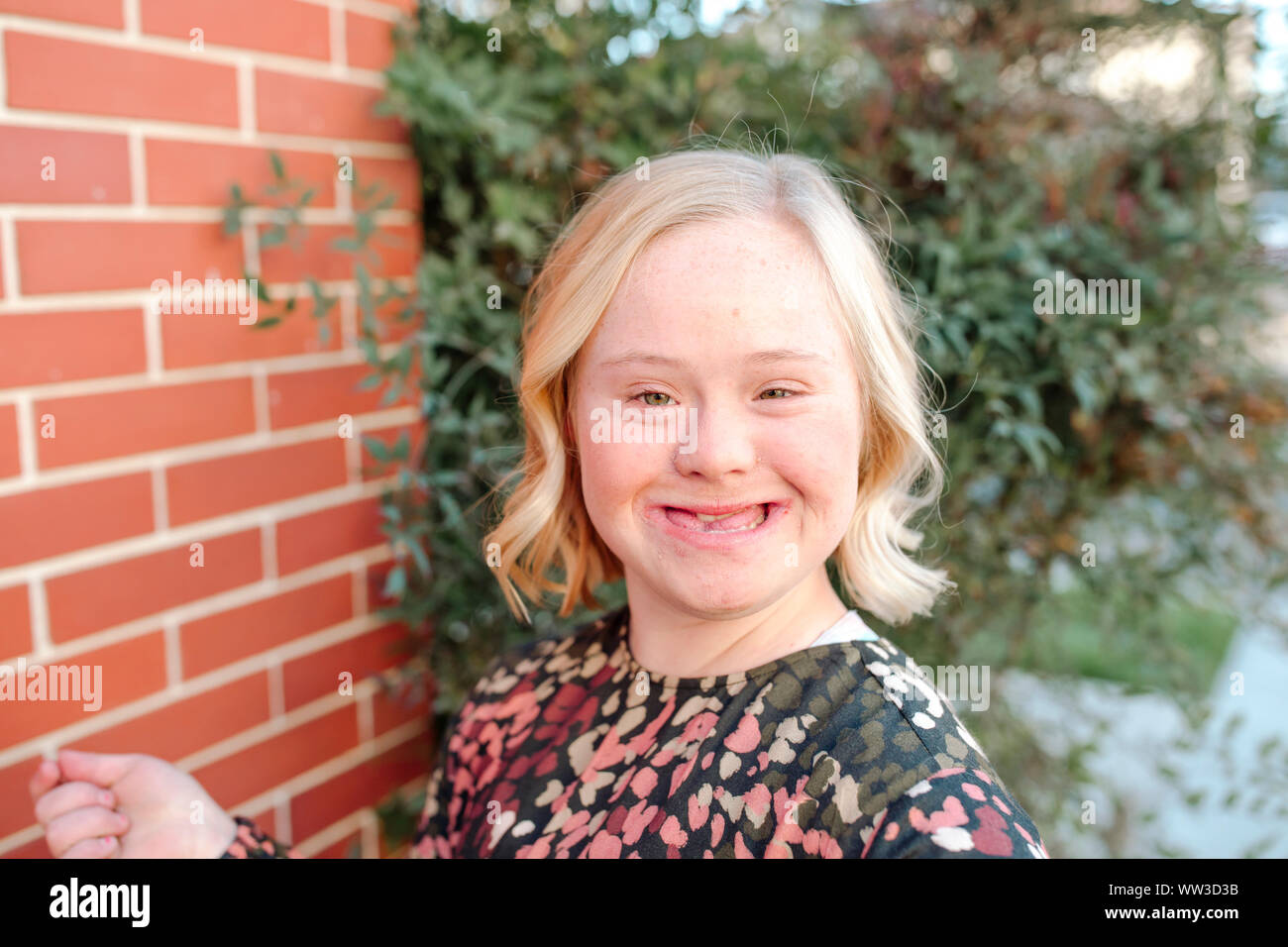 Happy teen girl with Down Syndrome smiling by brick wall on sunny day Stock Photo