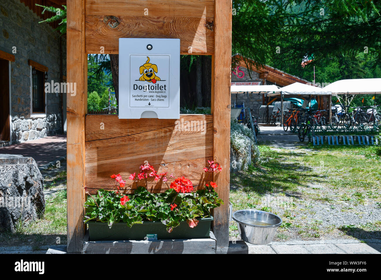 A wooden dog bag dispenser with a water bowl and a flowering plant in a mountain tourist park in summer, Courmayeur, Aosta, Italy Stock Photo