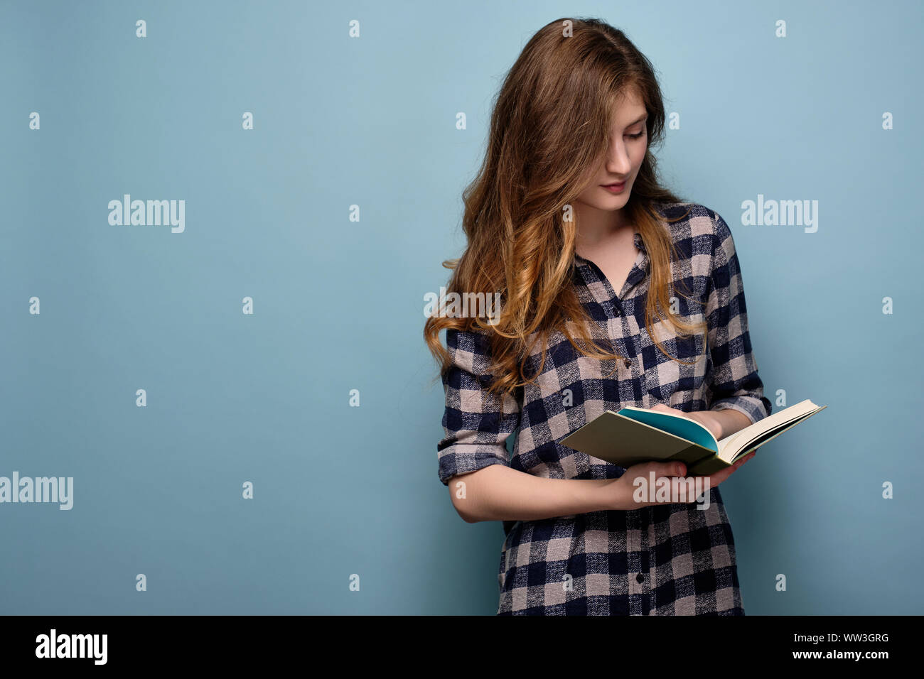 A young girl in a plaid shirt stands on a blue background and looks down in a book with his head bowed. Stock Photo