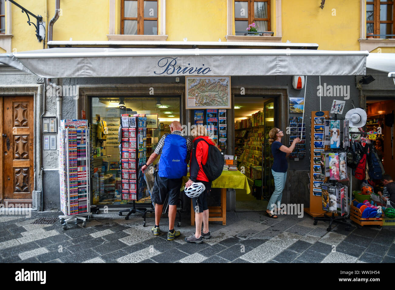 Exterior of a stationery shop with displays on the sidewalk and a couple of hikers looking at maps in the centre of Aosta, Aosta Valley, Alps, Italy Stock Photo
