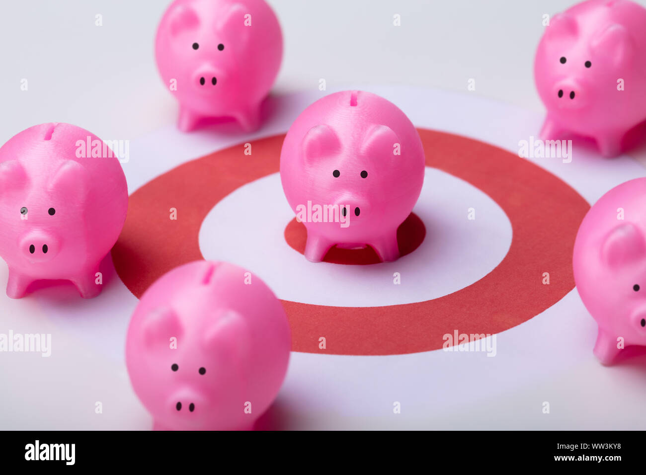 Pink Piggy Banks On Darts Target Surrounded With Other Piggy Banks Over White Desk Stock Photo