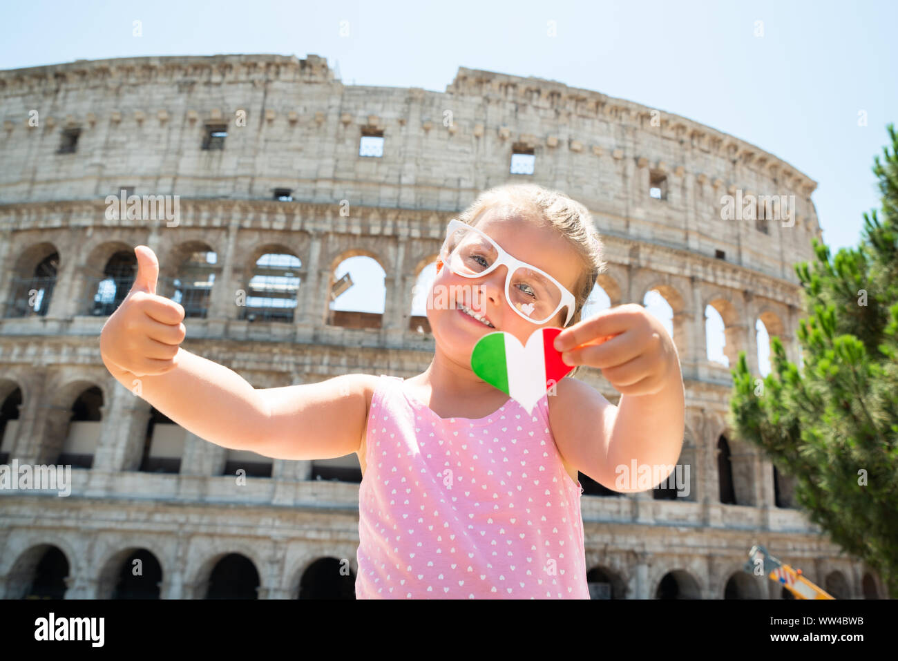 Girl Wearing Italian Flag Sunglasses, Holding Heart And Showing Thumb Up Near Colosseum, Rome, Italy Stock Photo