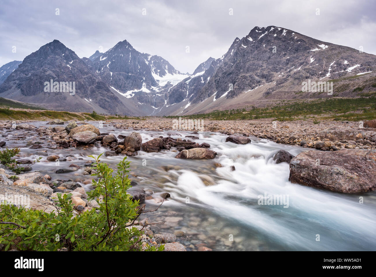 Wild Stream on fjell between rocks and mountains from Blåvatnet to sea, Lyngen alps in background and small green tree in foreground, Lyngen, Norway Stock Photo