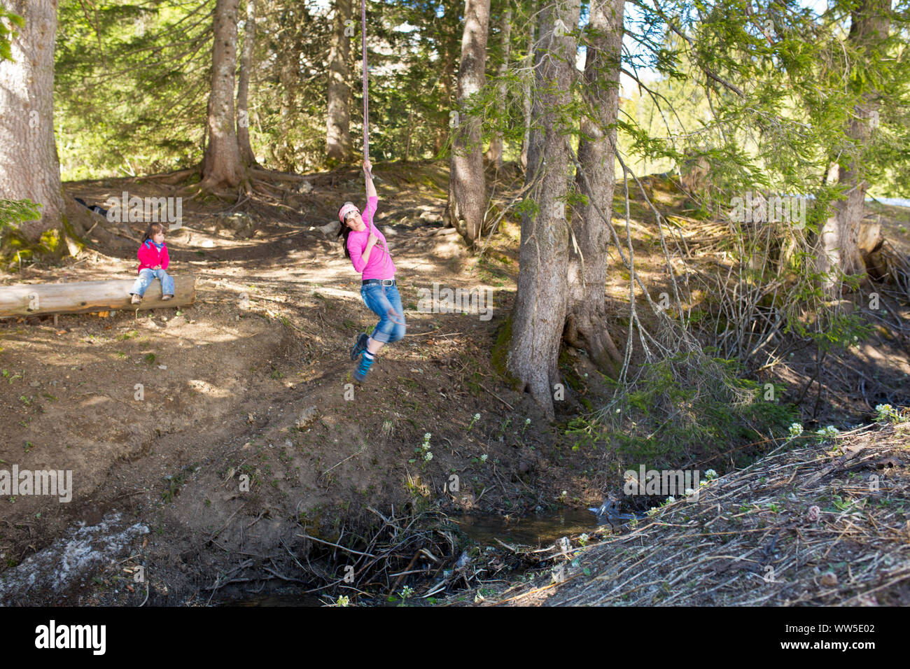 30-40 years old woman hanging on rope in the forest, swinging above a ditch, 5-year-old child in the background Stock Photo