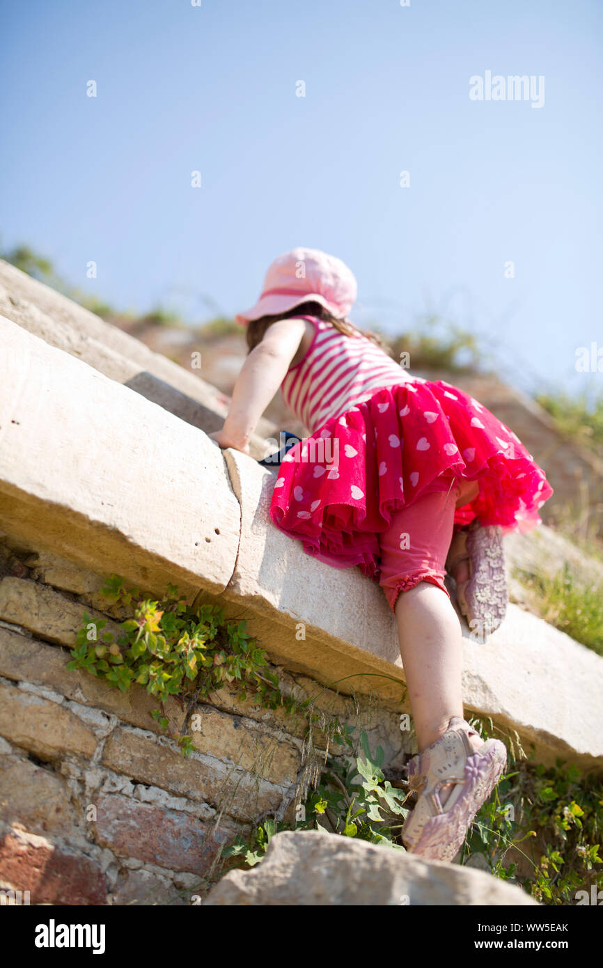 4-6 years old girl with red and white striped dress and pink hat climbing on wall Stock Photo
