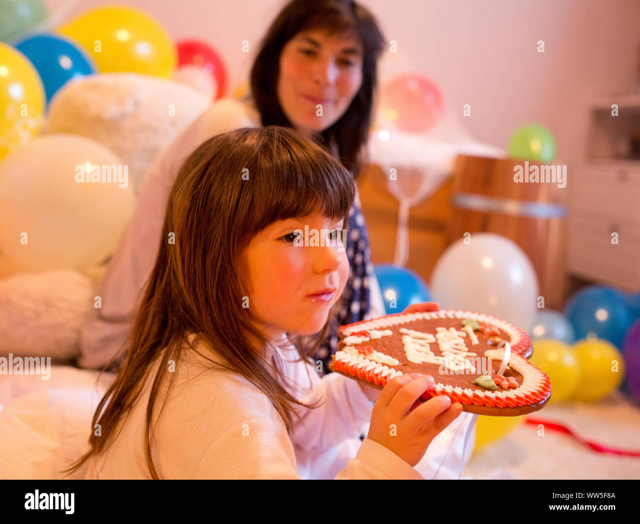 4-6 years old girl nibblings at a gingerbread heart in her room full of balloons, mother in the background Stock Photo