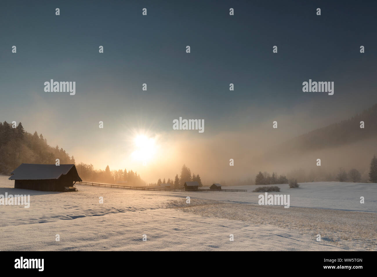 Hut in snow-covered alpine winter landscape close Garmisch Partenkirchen. In the background the rising sun in the morning fog with other wooden huts. Stock Photo