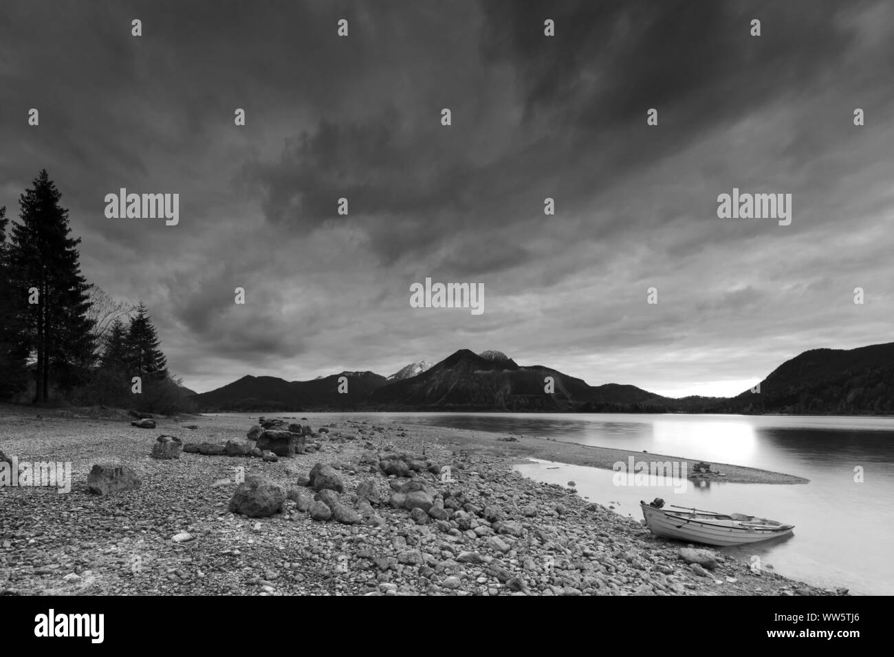 Rowing boat on the shore of the Walchensee shortly after sundown. In the background the Munich backyard mountains Herzogstand, Heimgarten and Jochberg and a peninsula from gravel, b/w Stock Photo
