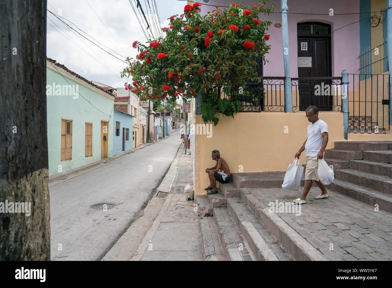 A man sitting on stairs in Santiago de Cuba, another walking down with purchases, houses, a balcony with flowers, a street building line with cables Stock Photo
