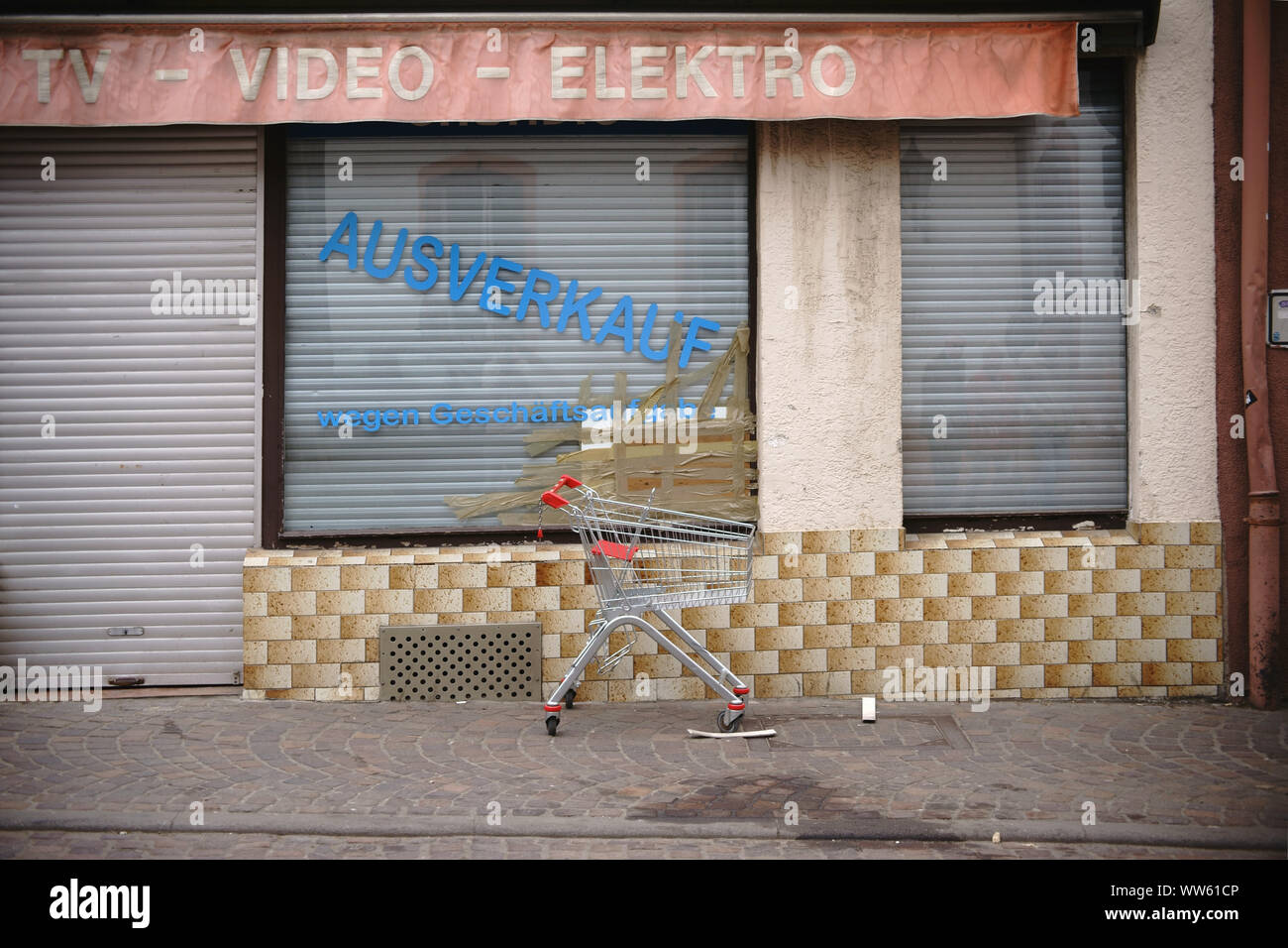 An empty shopping cart on the sidewalk in front of a closed electric goods shop with broken window pane, Stock Photo