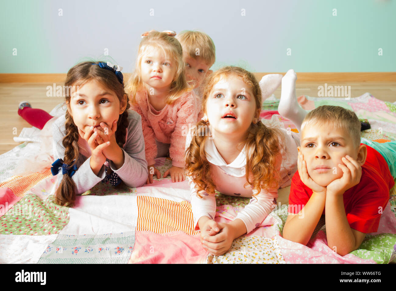 Close view of five adorable kids laying on a floor on a soft cover looking with curiousity on something behind the camera. Group of preschoolers watch Stock Photo