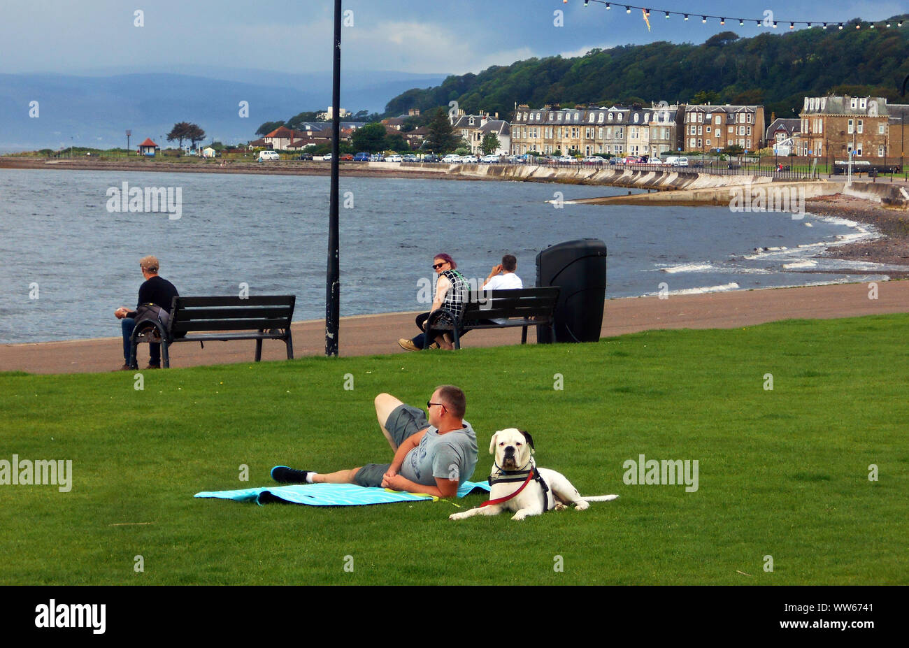 A few people sitting and relaxing at the sea front, holiday town, of Largs on the Firth of Clyde in Scotland. There is a man lying on the grass with a rather large black and white dog keeping its eye on things! Alan Wylie/ALAMY © Stock Photo