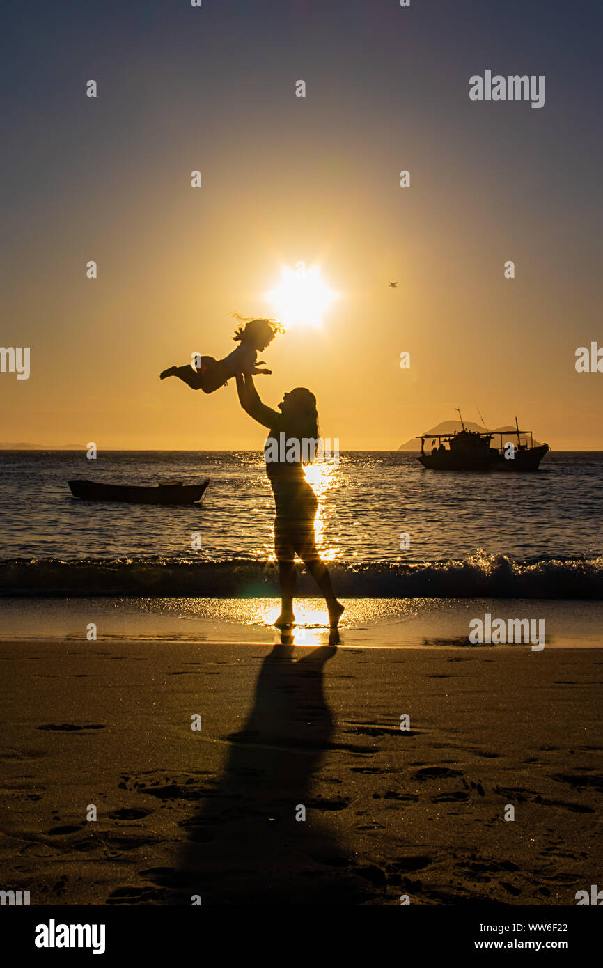 Silhouette of a woman standing on beach at sunset throwing her daughter in the air, Brazil Stock Photo
